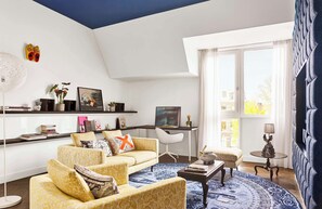 Un salon moderne avec un canapé, une table basse et un bureau avec une chaise. Une grande fenêtre avec des rideaux, une lampe et un vase décoratif ornent une table d’appoint.