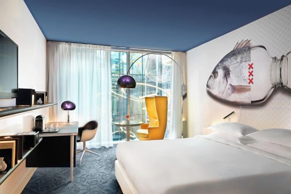 A hotel room with a bed, a desk, a chair, a window with curtains, and a wall-mounted fish artwork.
