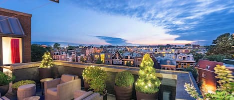 A rooftop terrace with wicker furniture, potted plants, and a city view at dusk.