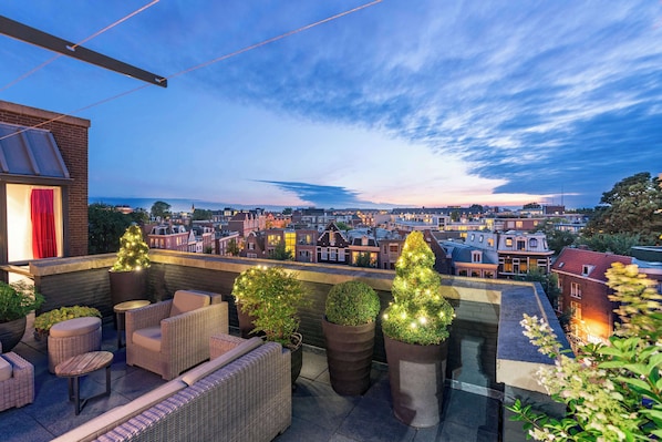 Terraza en la azotea con mobiliario de mimbre, plantas en macetas y vistas a la ciudad al atardecer.