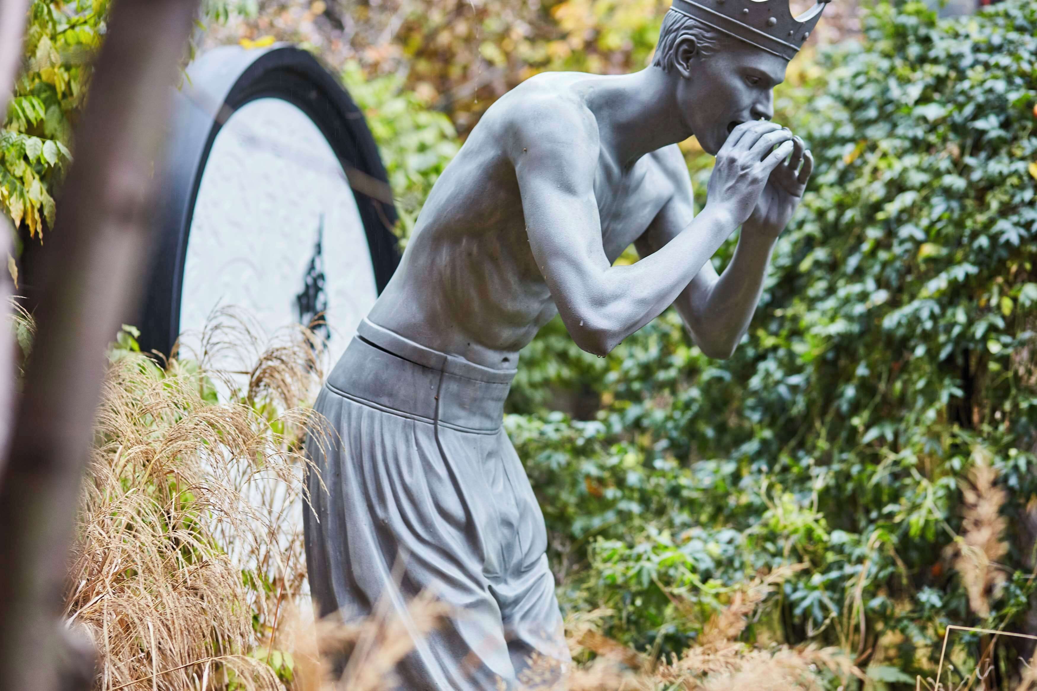 A statue of a muscular figure in a gray outfit with a crown, standing in a natural setting with greenery and a surfboard in the background.