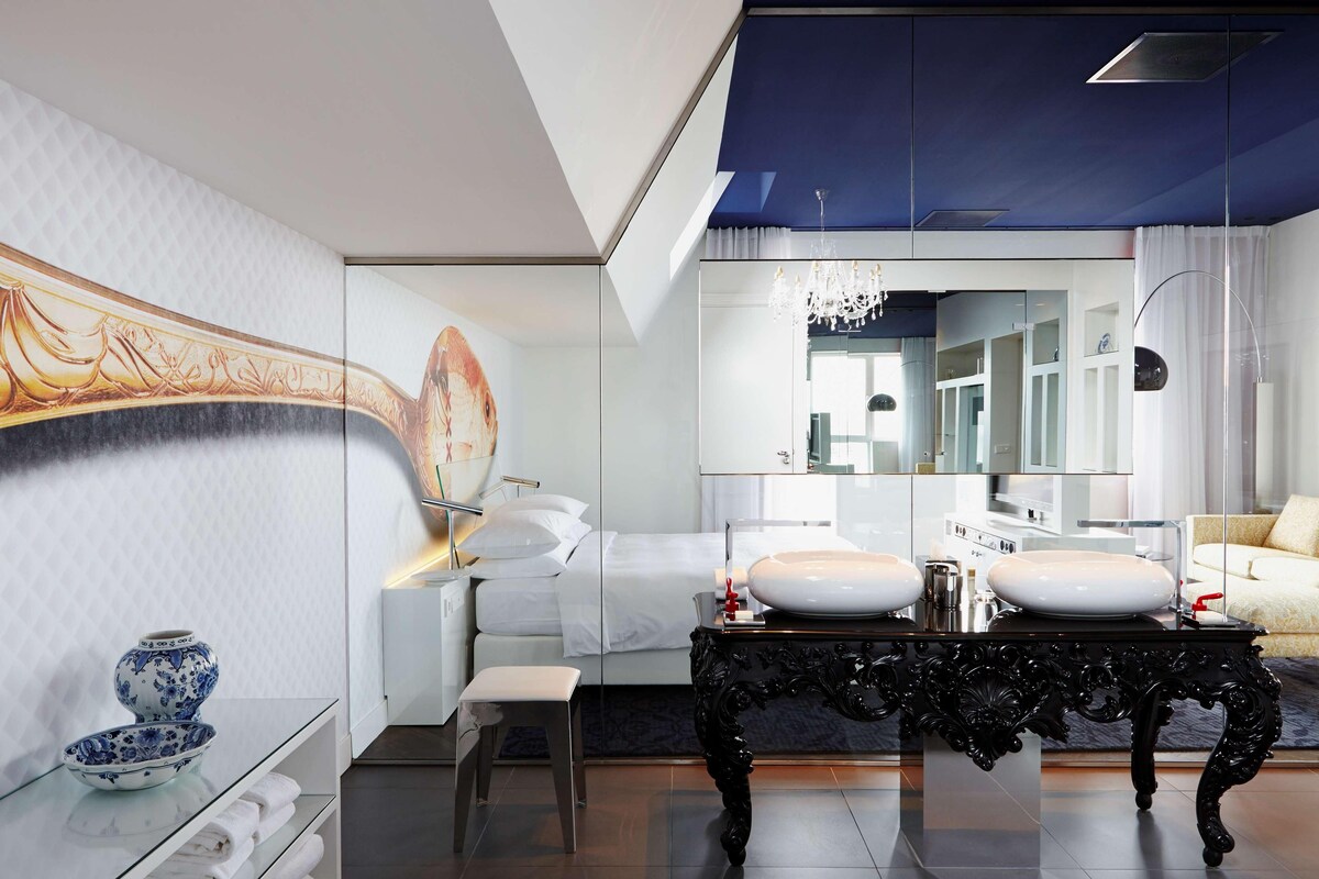 A modern bathroom with a large mirror, two sinks, a vanity with a decorative bowl, and a bed with white linens.