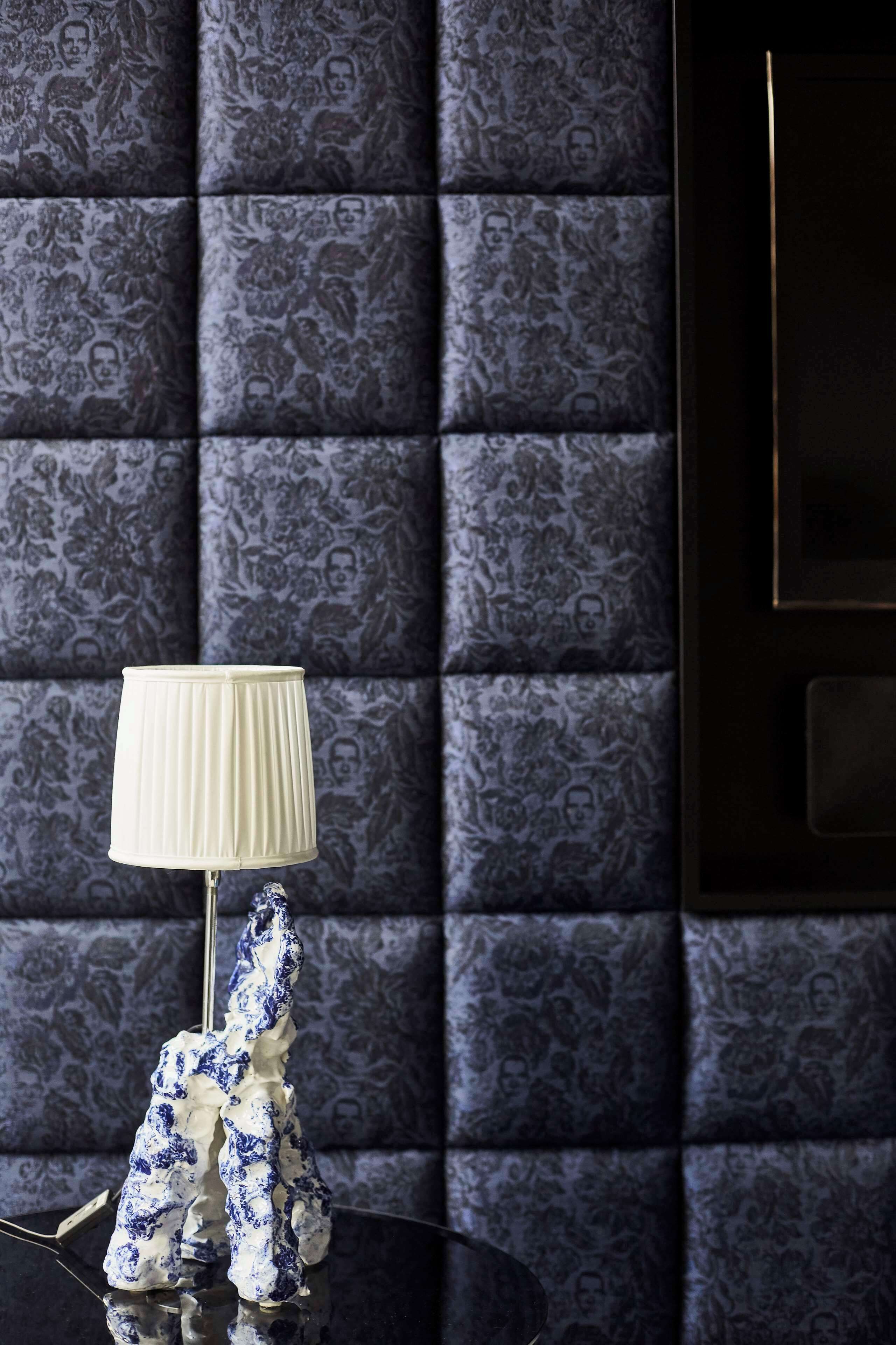Een kamer met een donkere, patroonbezetting aan de wand, een witte lamp en een blauw-wit beeld op een reflecterend oppervlak.