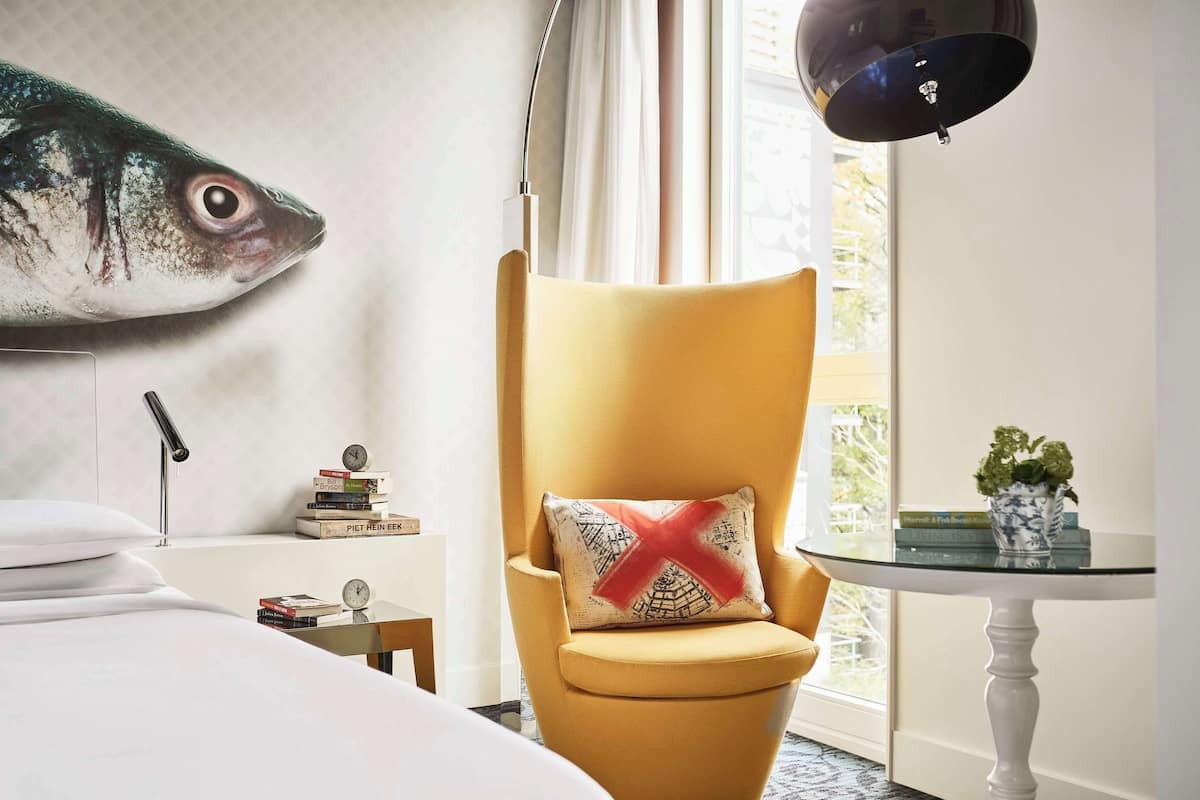 Habitación de hotel con un gran mural de peces, un sillón amarillo con un cojín, una mesita de noche con libros y un reloj, y una ventana con vistas a zonas verdes.