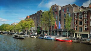 A canal with boats, a row of buildings with flags, and bicycles parked along the sidewalk.
