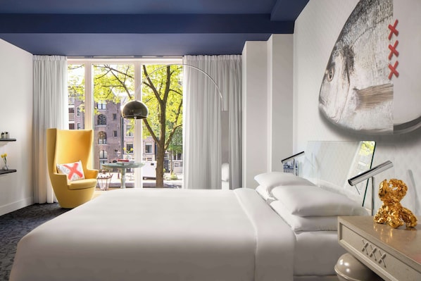 Habitación de hotel con una cama grande, un sillón amarillo, vistas de la ciudad a través de la ventana y una obra de arte colgada en la pared.