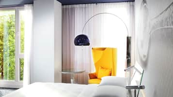Una habitación de hotel con una cama, un sillón amarillo, una mesa de vidrio, una lámpara de pie moderna y un ventanal con cortinas.