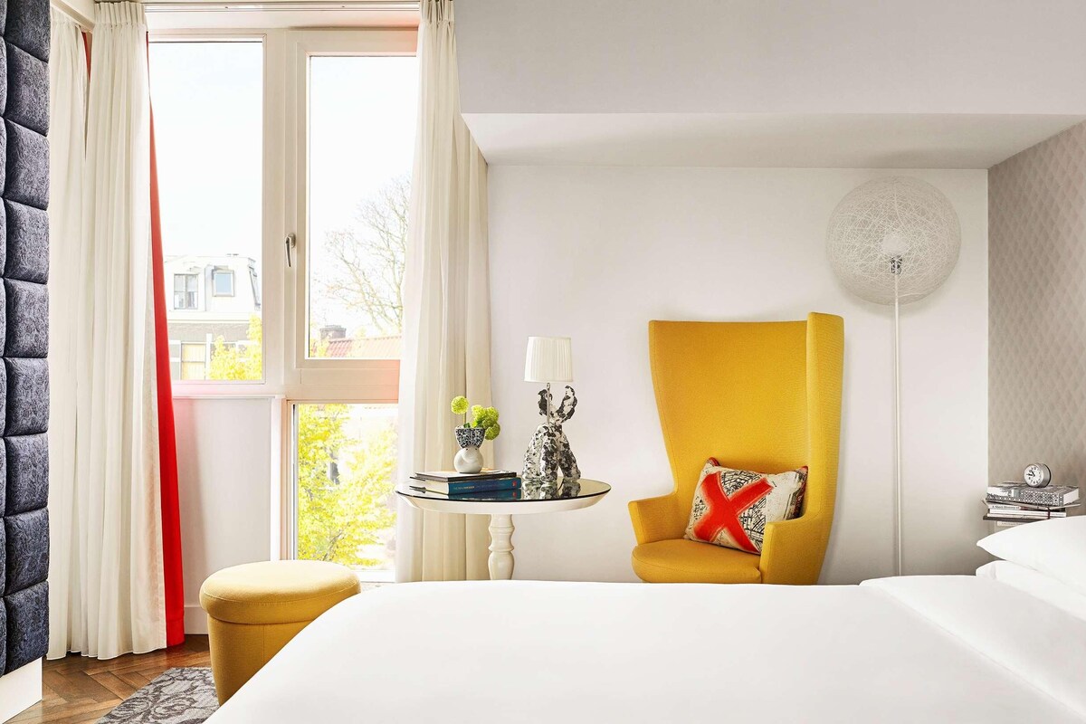 A hotel room with a bed, a yellow armchair, a round table with a lamp, and a large fish painting on the wall.