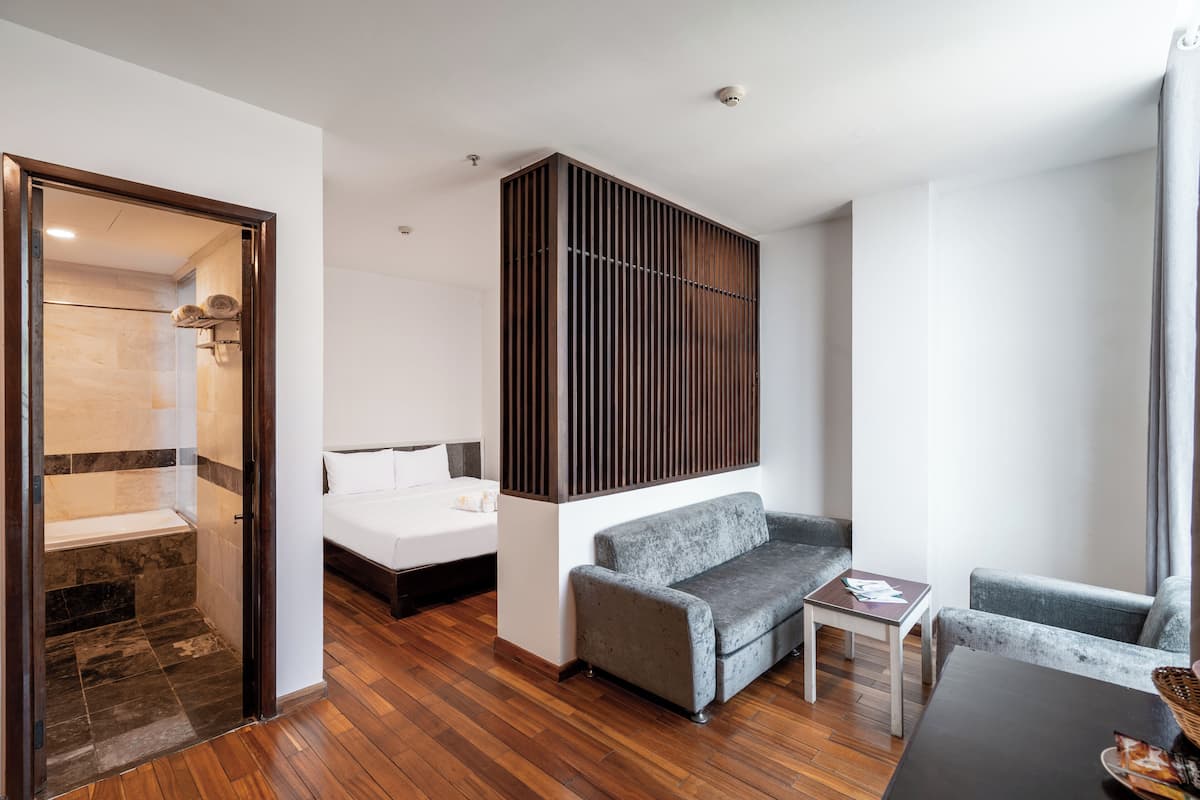 Una habitación de hotel moderna con suelo de madera, un sofá gris, una mesita, una cama y zona de ducha.