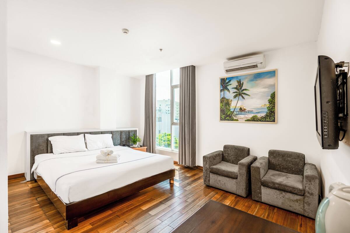 Una habitación de hotel con una cama grande, un televisor, un cuadro de una playa y dos sillones.