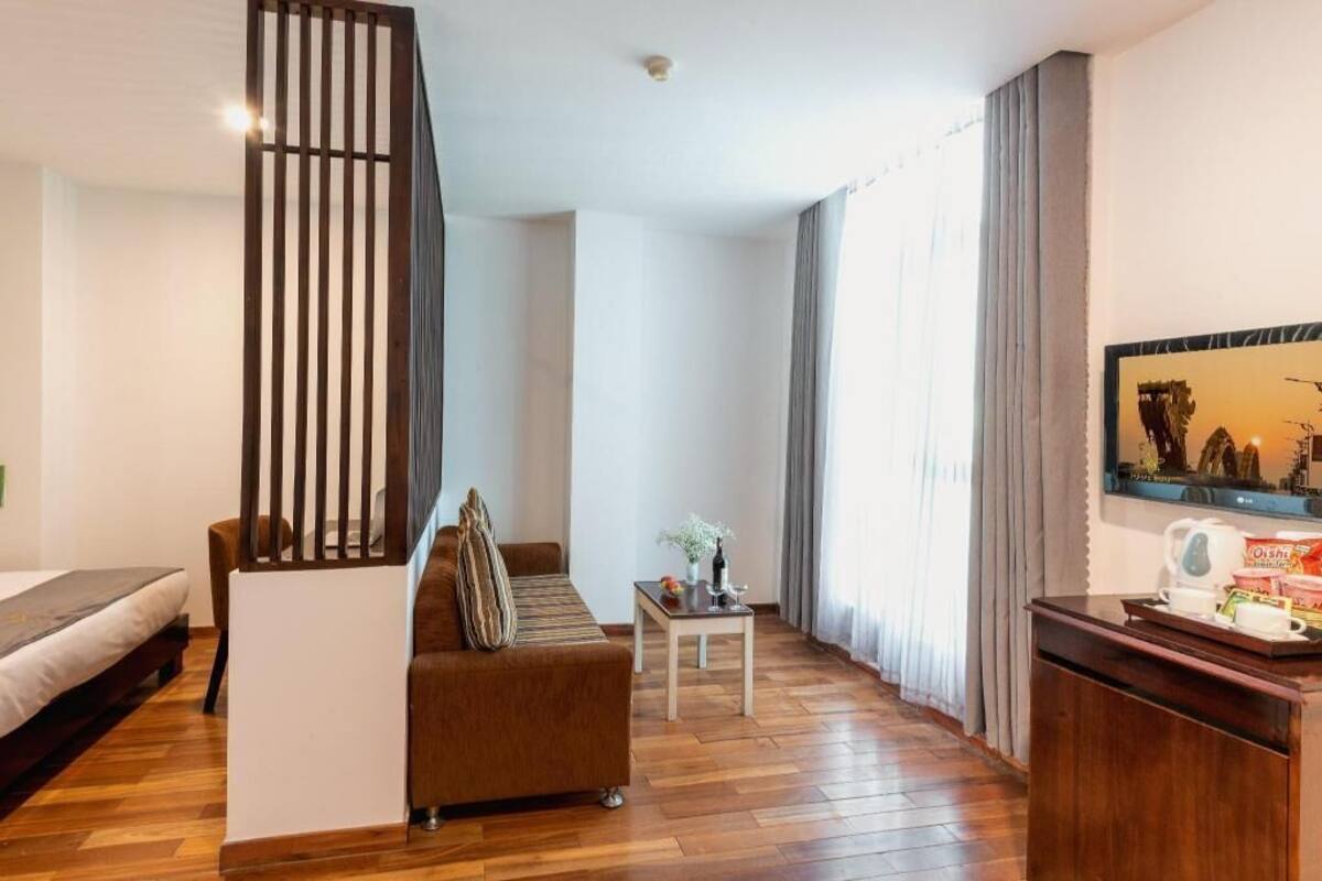 Una habitación con piso de madera, un sofá, una pequeña mesa con un florero, una cama, un escritorio con una bandeja y un cuadro en la pared.