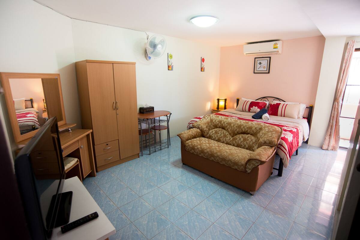 A compact hotel room with a bed, a sofa, a desk, a wardrobe, and a fan.