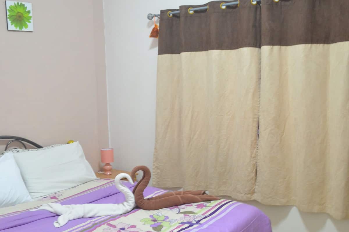 A hotel room with a bed, a wall-mounted air conditioner, a curtain, and a small table with a pink lamp.