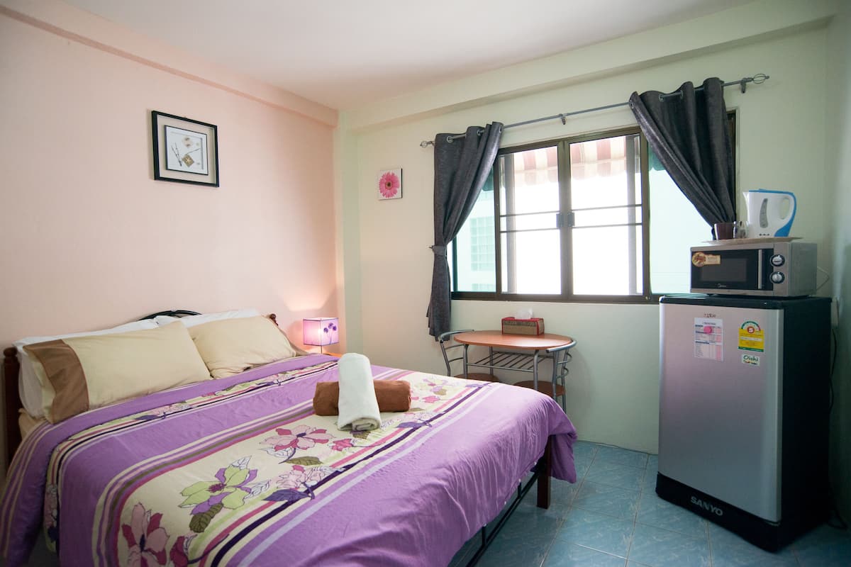 A bedroom with a bed, a small table, a refrigerator, and a window with curtains.