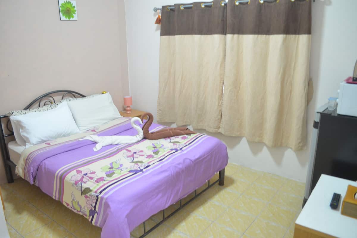 A bed with a purple bedspread, a white pillow, and a brown towel. There is a nightstand with a lamp and a remote control on it. A curtain is partially drawn, and a small plant is on the wall.