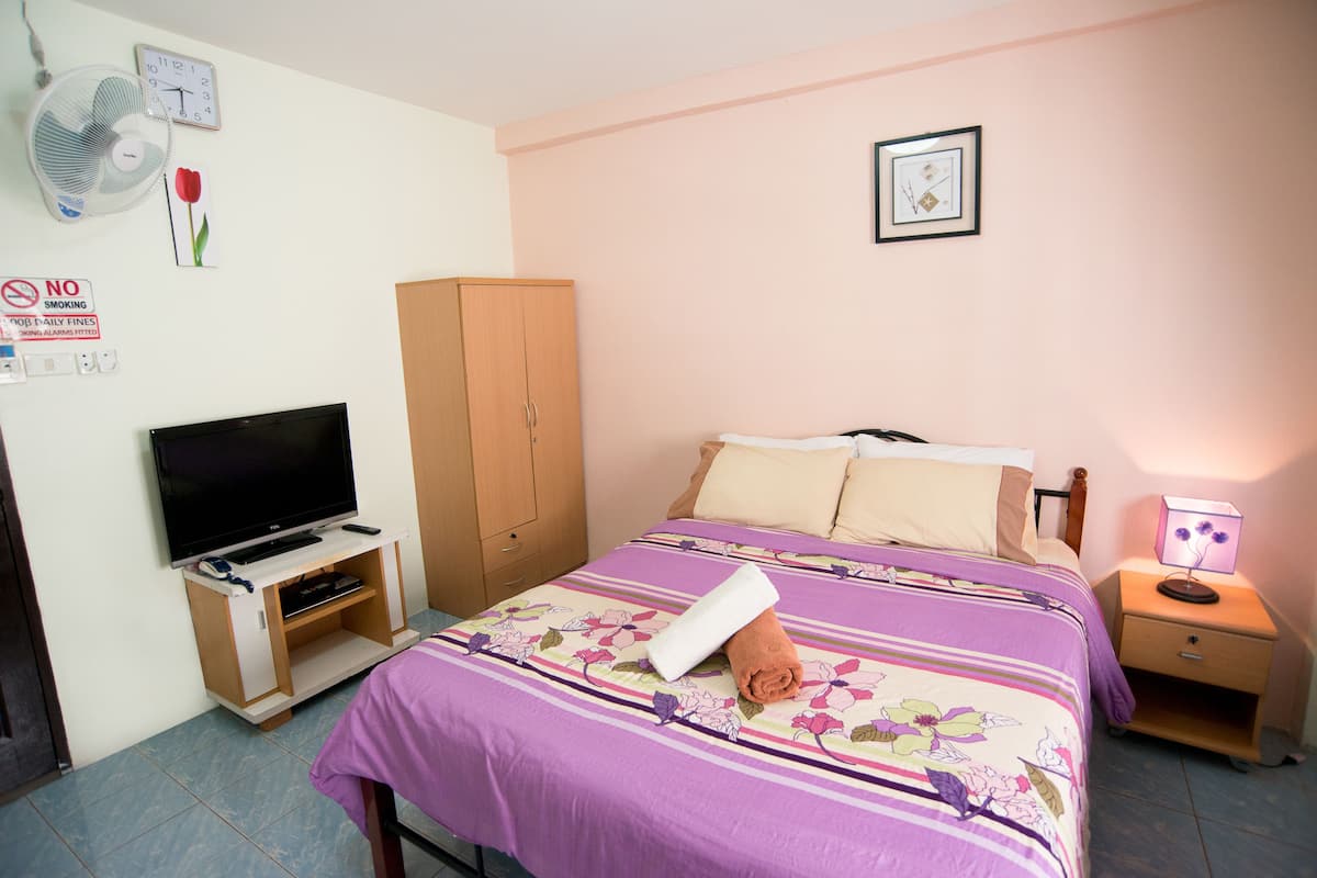 A bedroom with a bed, a TV, a wardrobe, a nightstand, a lamp, a fan, and a clock on the wall.