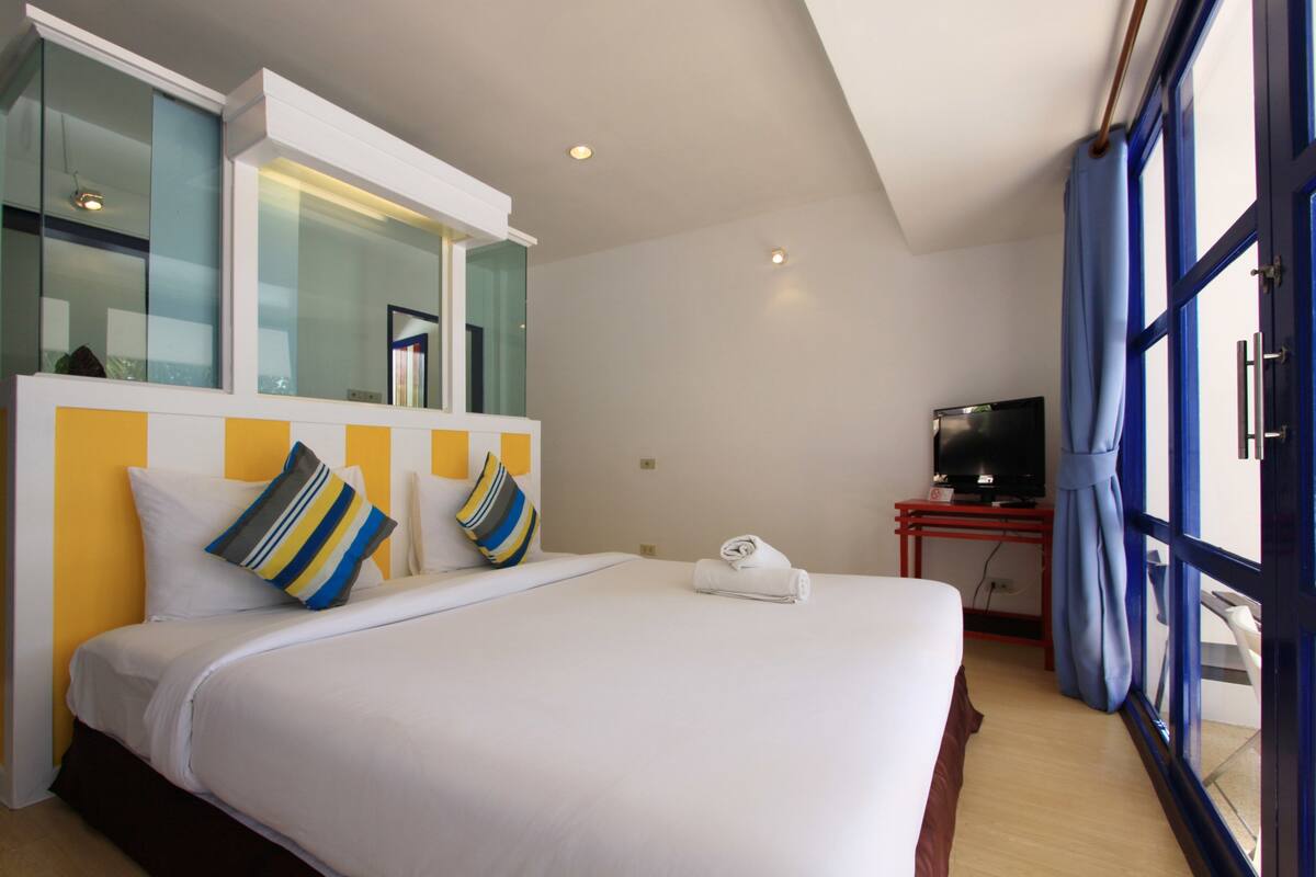 A hotel room with a large bed, a TV on a stand, a window with curtains, and a small table with a remote control.