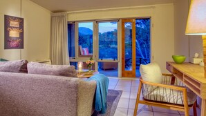 Ein gemütliches Wohnzimmer mit einer Couch, einem Holzstuhl, einem Tisch mit einer Schüssel und Blick auf eine gebirgige Landschaft durch große Schiebeglastüren.