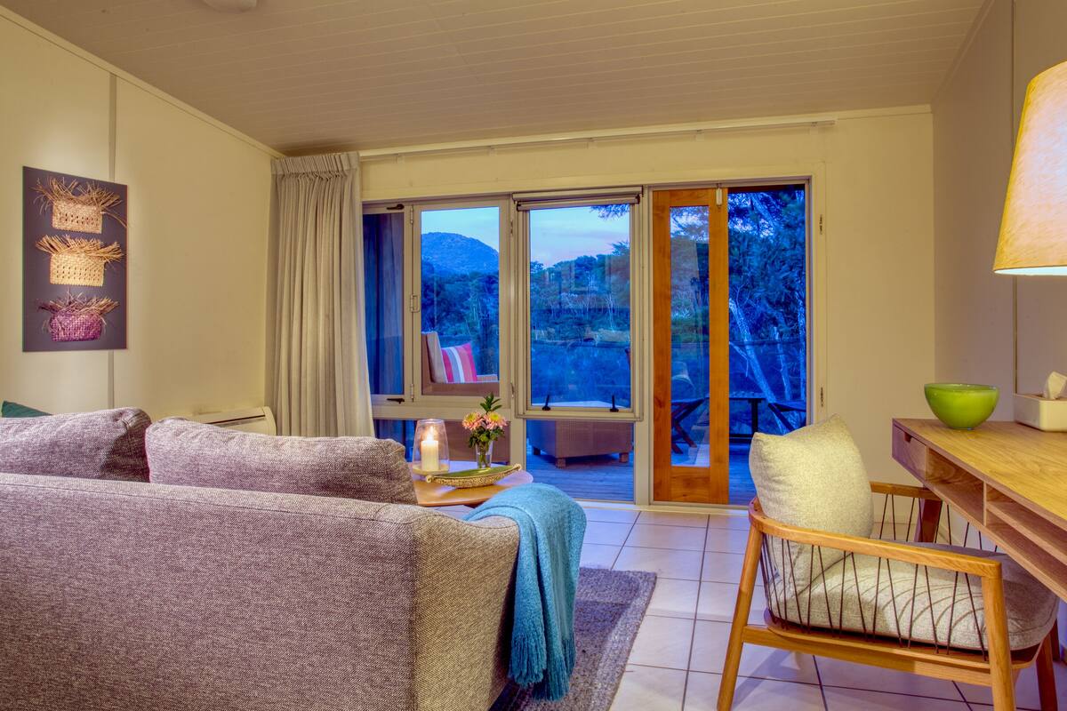 A cozy living room with a sofa, a wooden chair, a table with a bowl, and a view of a mountainous landscape through large sliding glass doors.