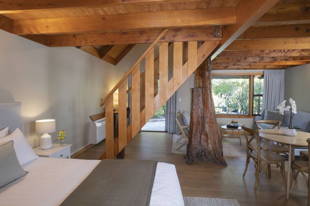 A bedroom with a wooden staircase, a bed, a nightstand, a lamp, and a window with a view of the outdoors.
