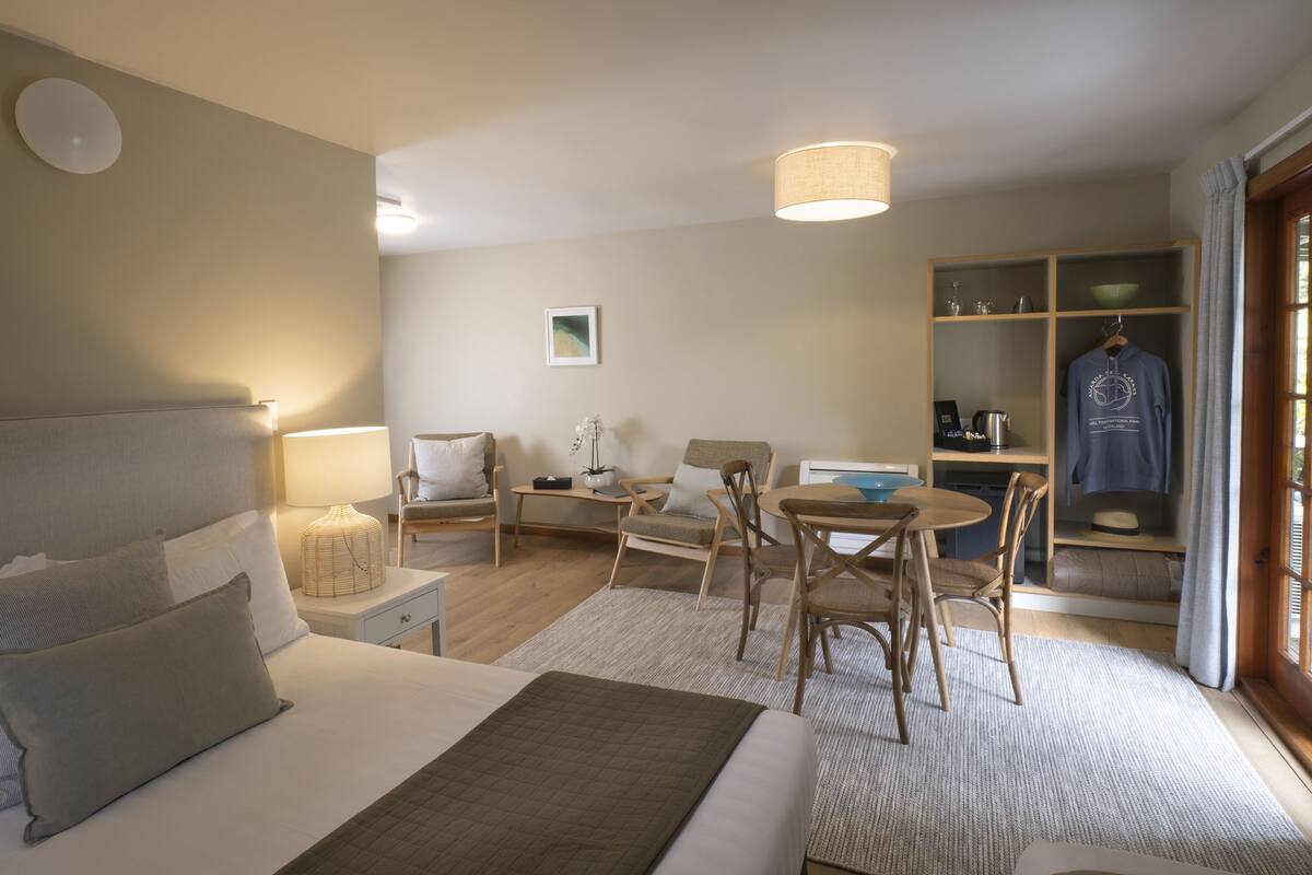 A hotel room with a bed, a desk, a chair, a small table, a bookshelf, and a hanging garment.