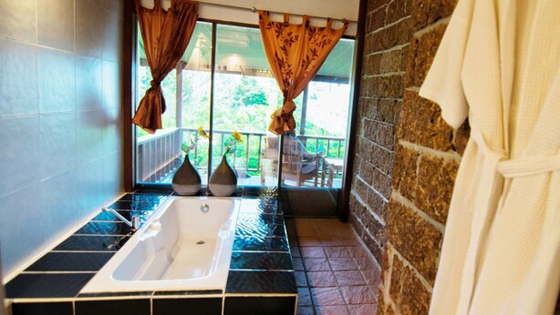 A bathroom with a bathtub, a window with curtains, a brick wall, and a white robe hanging on the wall. - Faye Orchid Resort (Ko Chang)