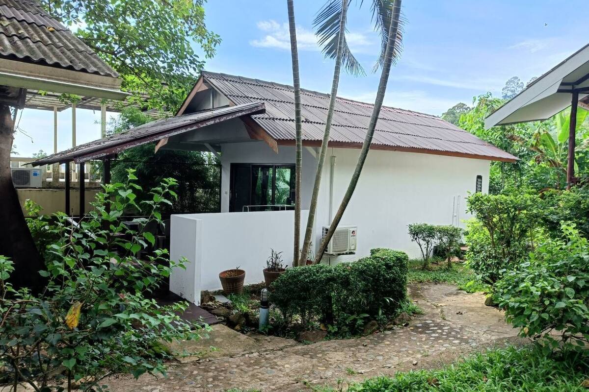 A white house with a tiled roof, surrounded by lush greenery and a stone pathway.