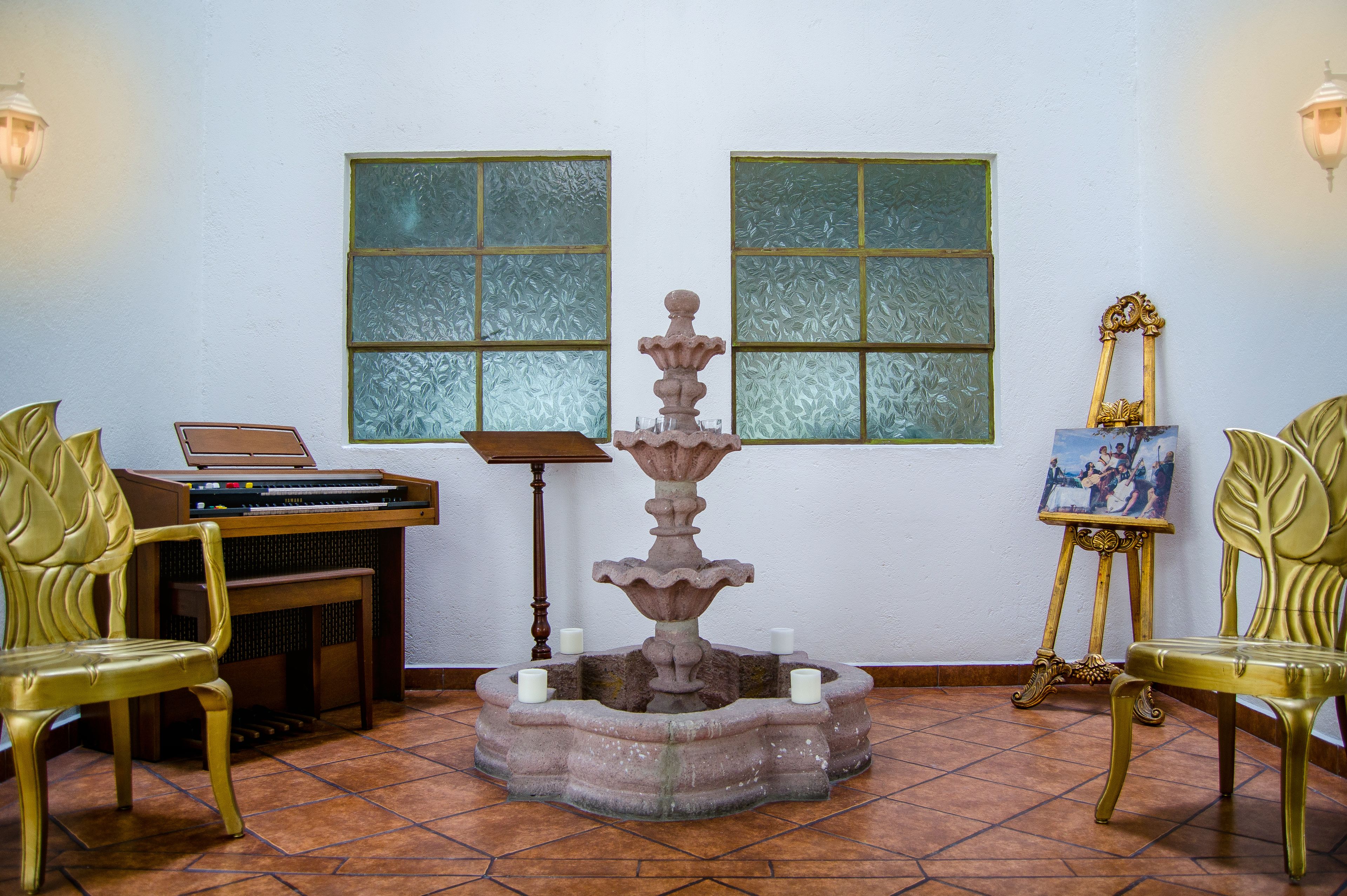 A room with a stone fountain, two green chairs, a wooden table with a keyboard, and a painting on an easel.