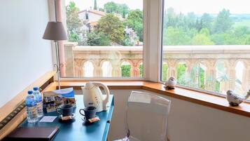 Una habitación con una mesa dispuesta con una tetera, tazas y botellas de agua, una silla transparente y vistas a un balcón con barandilla y vegetación.