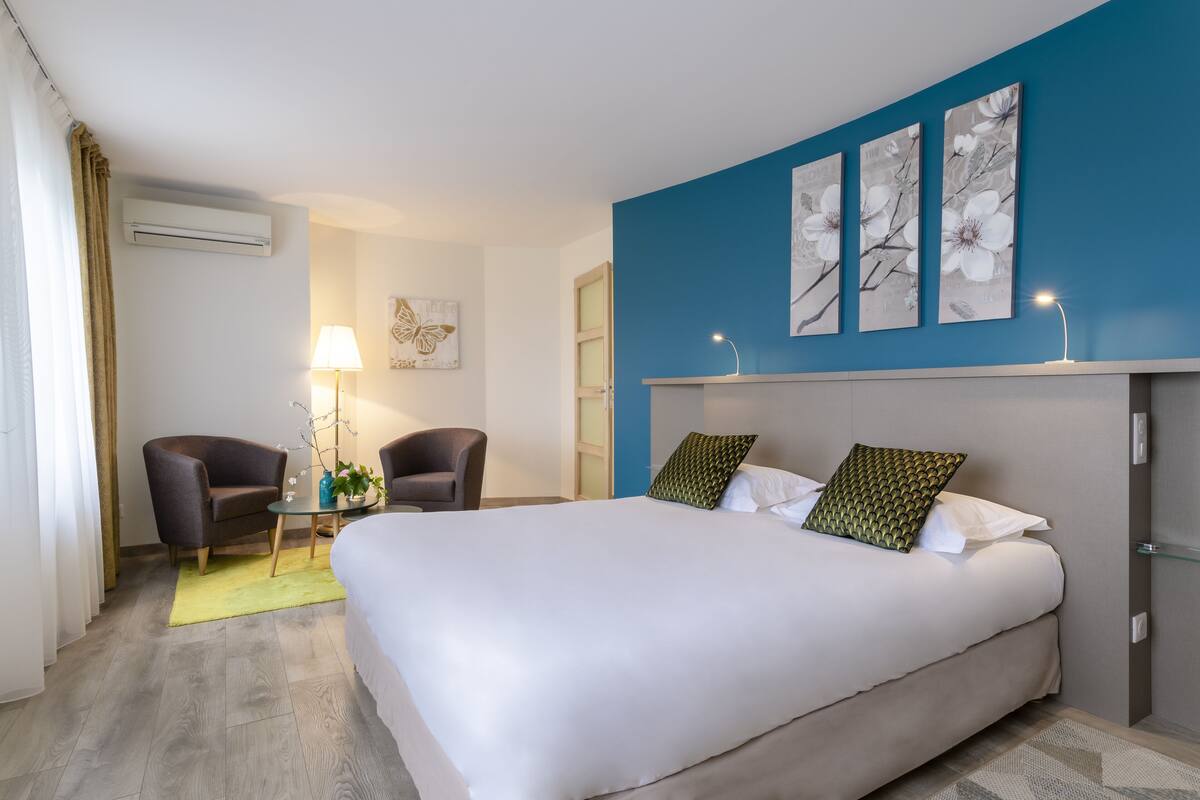 A hotel room with a large bed, two armchairs, a small table with a lamp, and artwork on the walls.