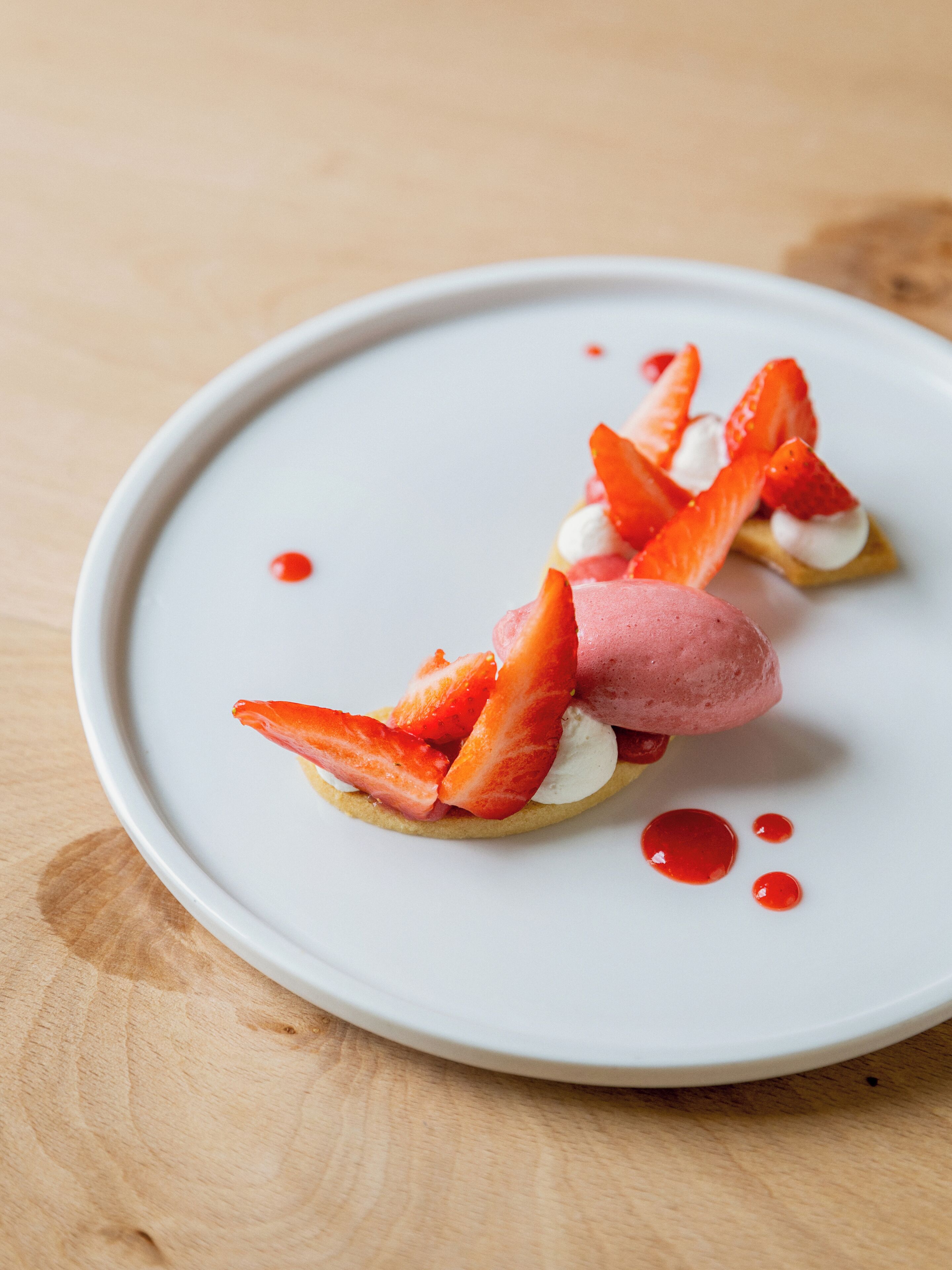 Un postre sobre un plato blanco, con fresas y una porción rosada, posiblemente helado, sobre una mesa de madera.