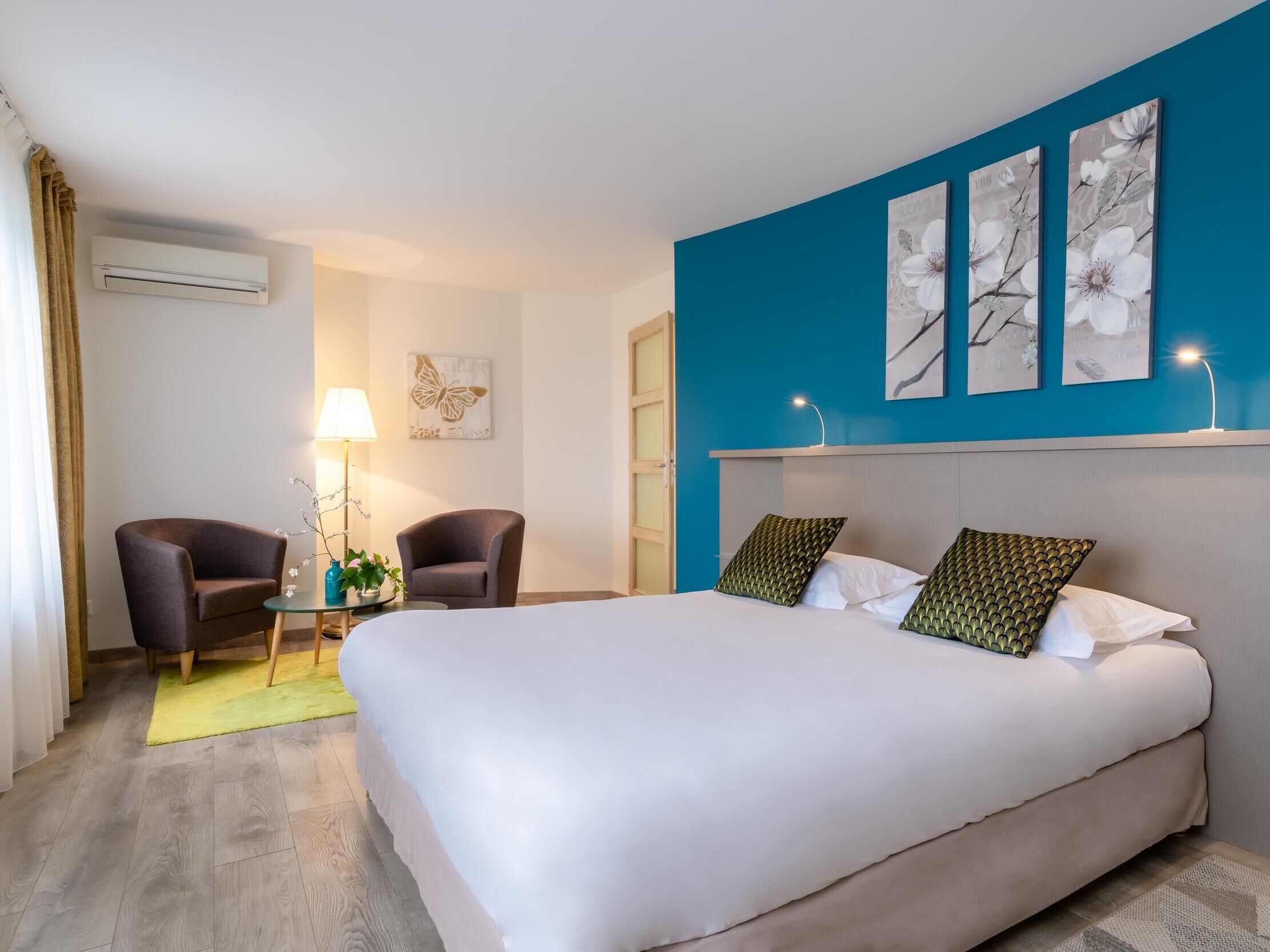 A hotel room with a bed, two armchairs, a small table with a lamp, and a wall-mounted air conditioner.