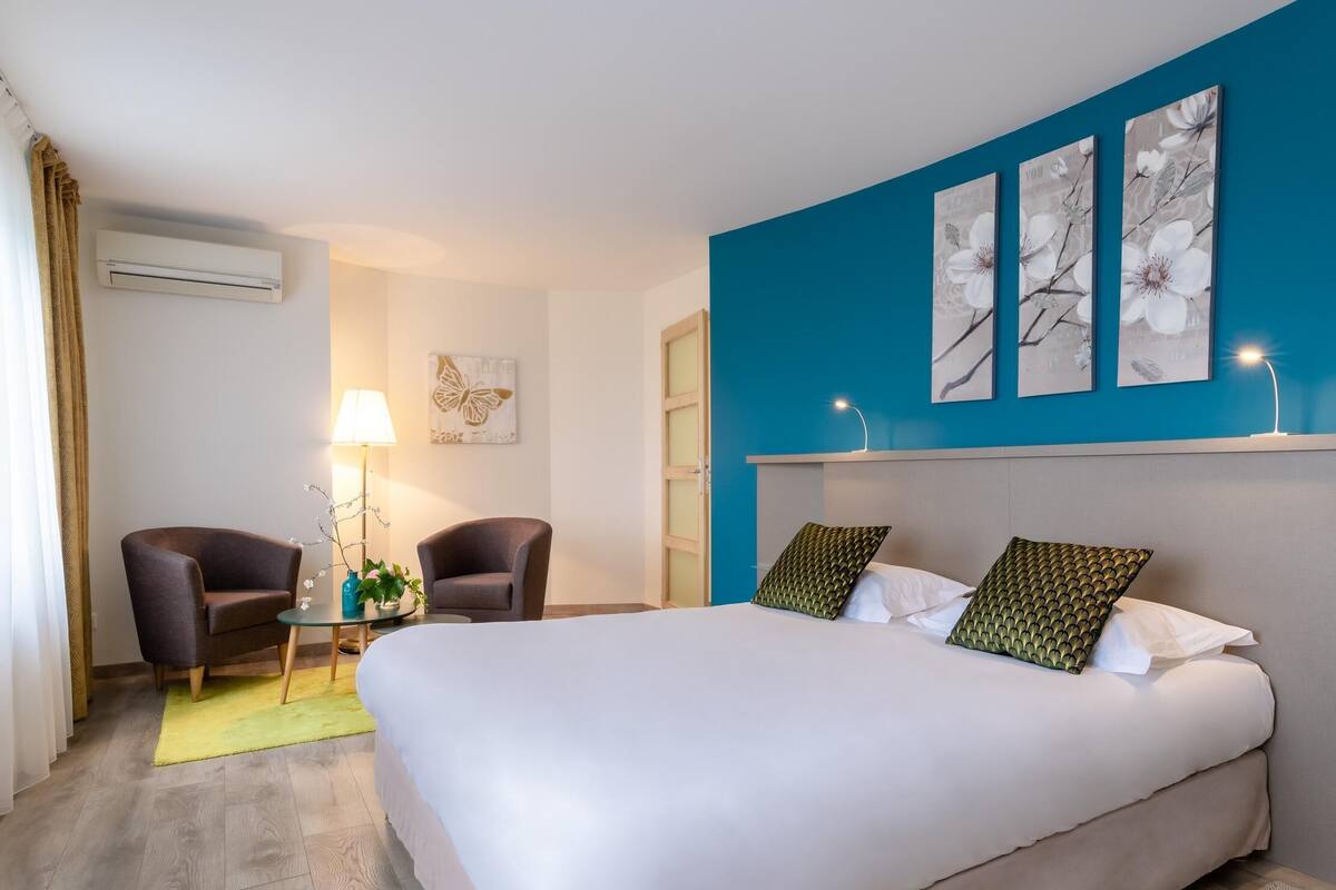 A hotel room with a bed, two armchairs, a small table with a lamp, and a wall-mounted air conditioner.
