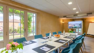 A conference room with a long table, blue chairs, a projector screen, and a view of the outdoors through large windows.