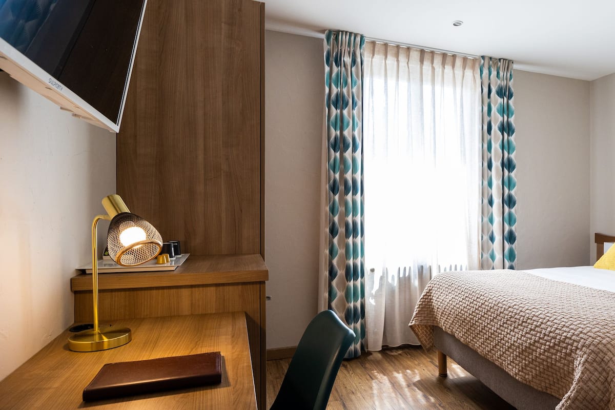 A hotel room with a bed, a desk, a chair, a lamp, and two framed pictures on the wall.