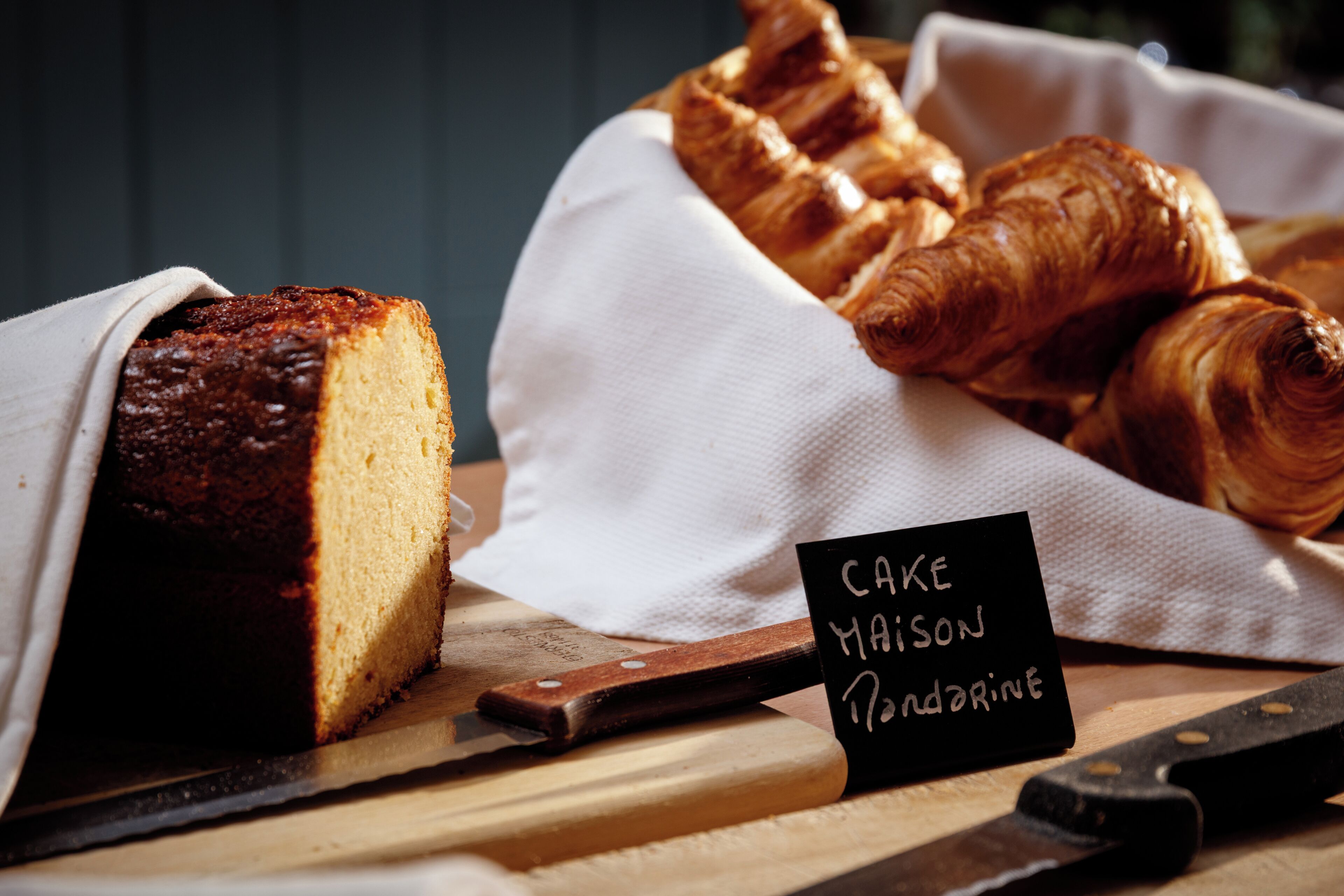 Une miche de gâteau et des croissants sur une planche en bois, accompagnés d’un couteau et d’une pancarte indiquant « Gâteau Madison ».