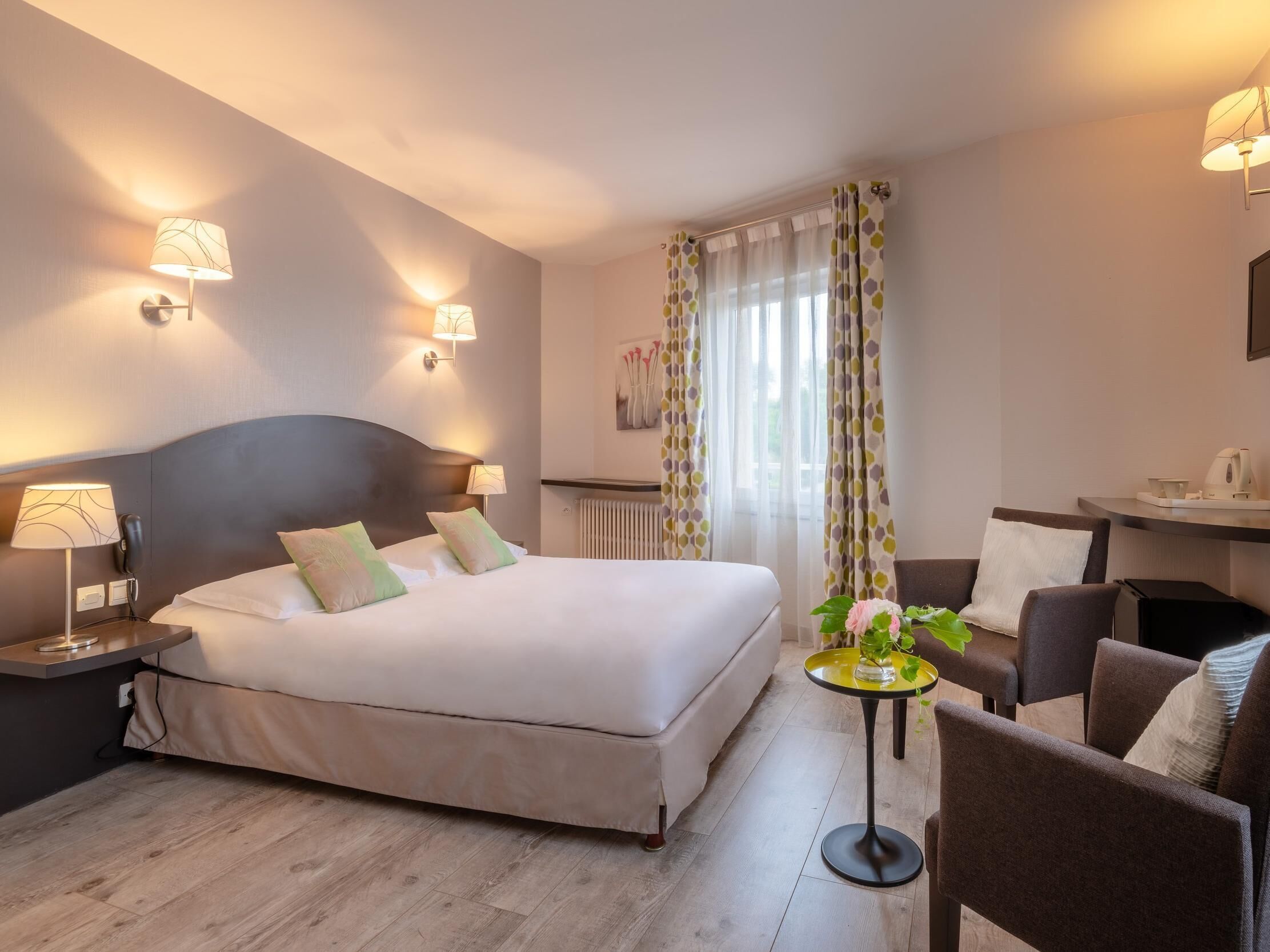 A hotel room with a bed, bedside lamps, a small table with a plant, a chair, and a desk with a kettle.