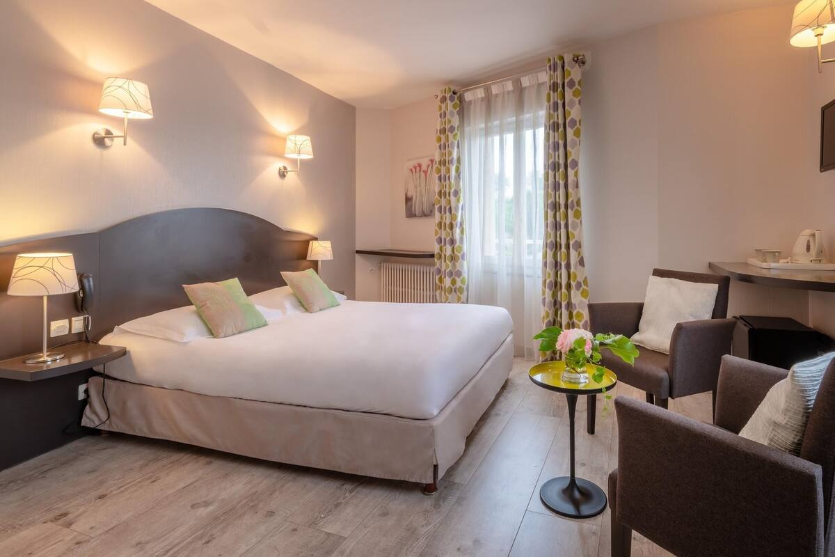 A hotel room with a bed, bedside lamps, a small table with a plant, a chair, and a desk with a kettle.