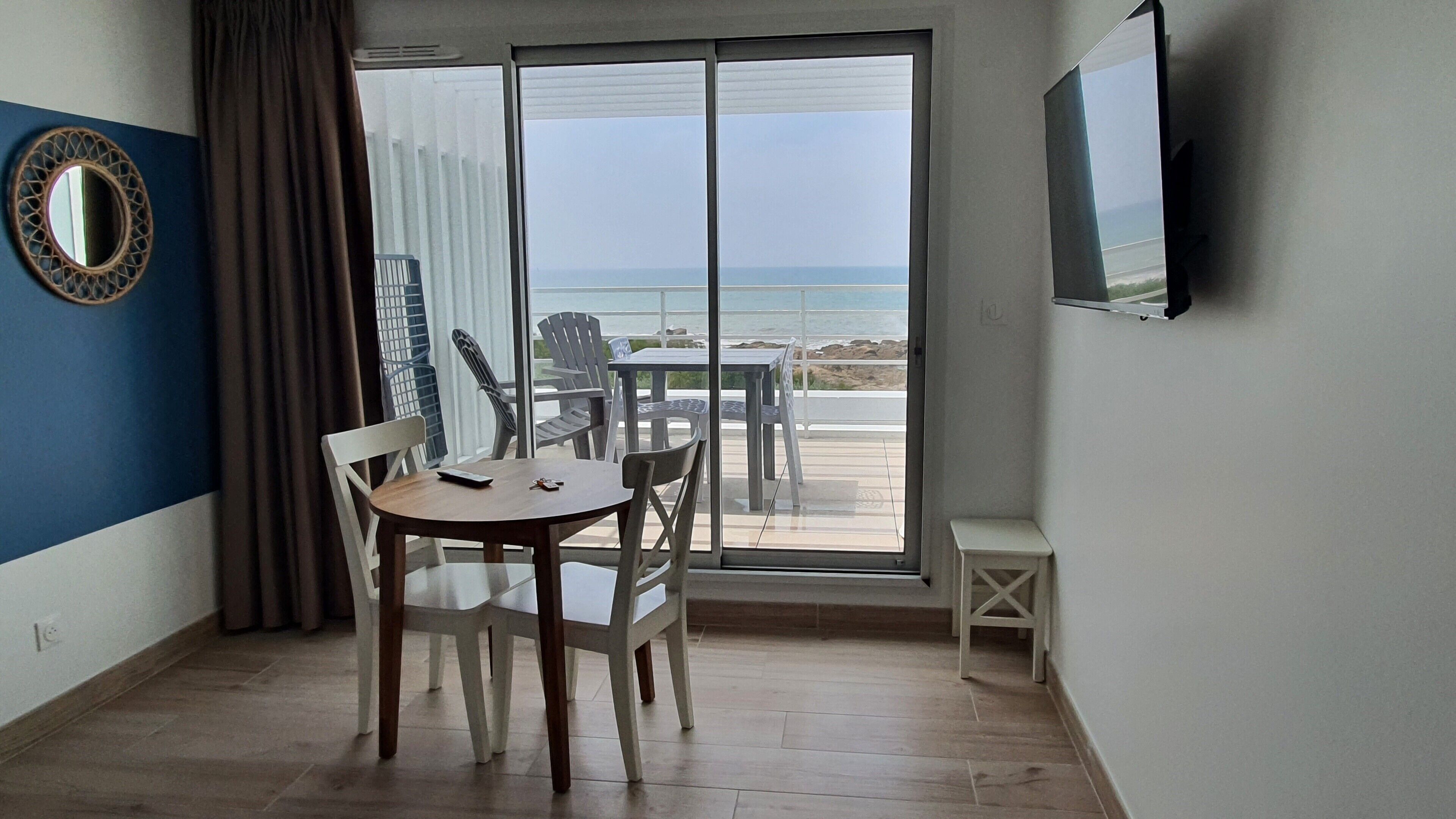 Habitación con un gran ventanal corredero que da a un balcón con vista a la playa. Hay un espejo redondo en la pared, una pequeña mesa con sillas y un televisor montado en la pared.