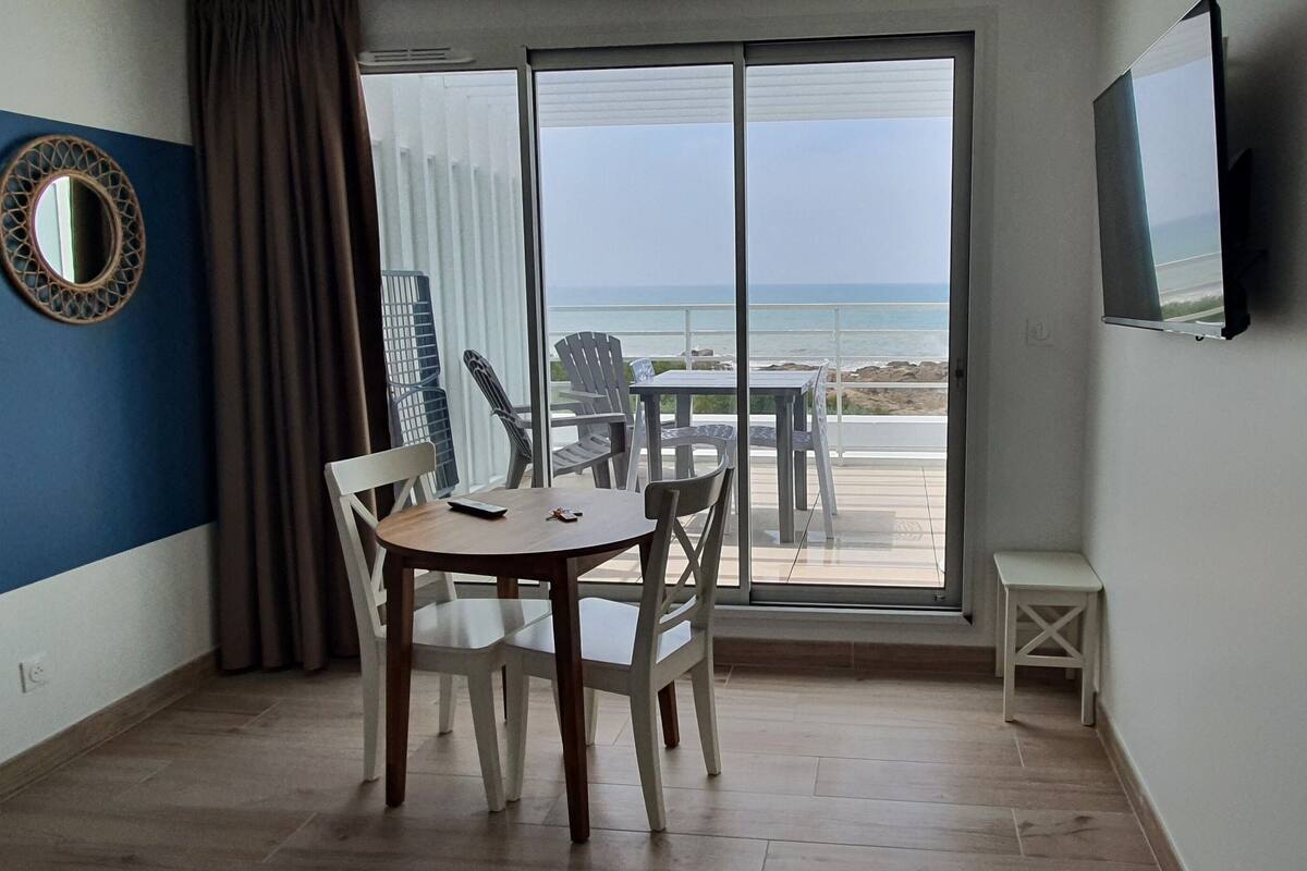 Une pièce dotée d’une grande porte-fenêtre coulissante donnant sur un balcon avec vue sur la plage. On y trouve un grand miroir rond accroché au mur, une petite table avec des chaises et un téléviseur fixé au mur.