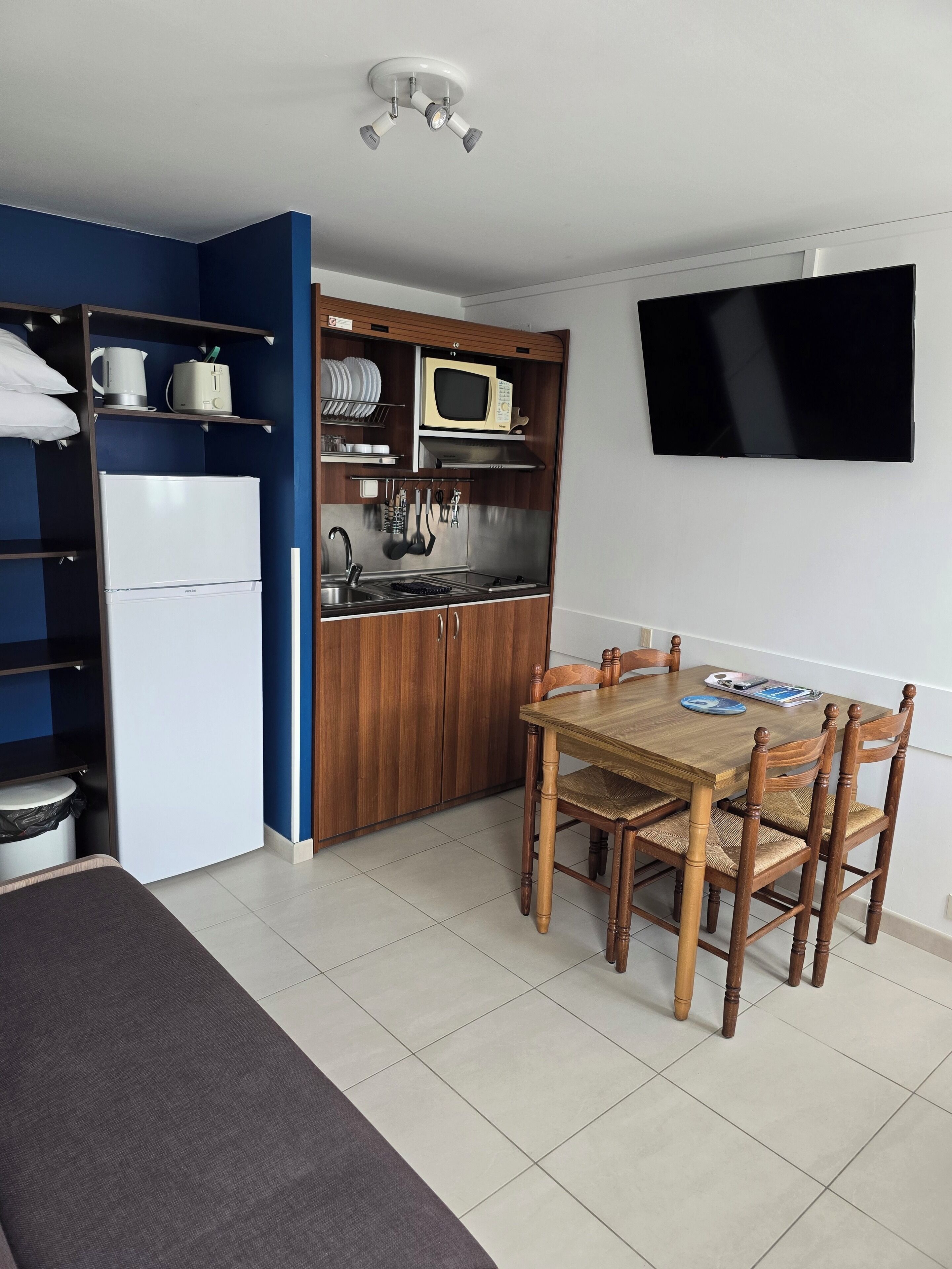 Cocina compacta con microondas, estufa y lavabo, una mesa de comedor con sillas y un televisor montado en la pared.