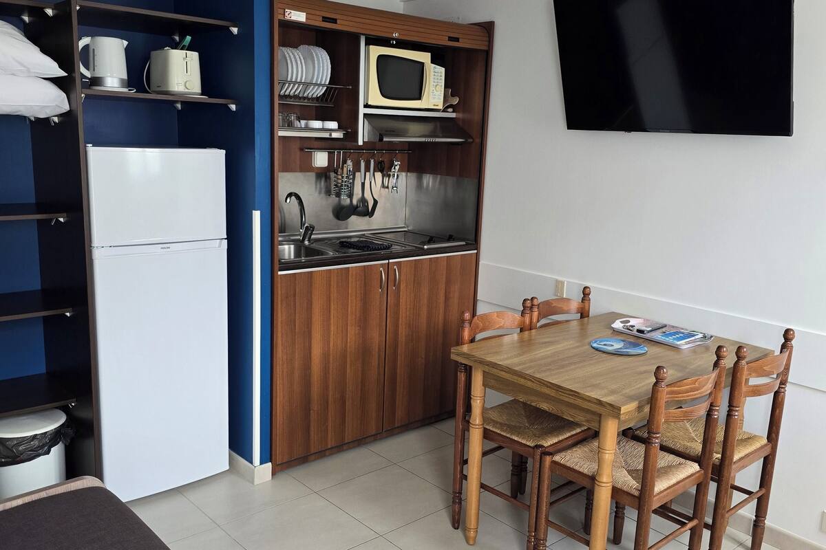 Une cuisine compacte équipée d’un four à micro-ondes, d’une cuisinière et d’un évier, ainsi qu’une table à manger avec des chaises et un téléviseur fixé au mur.