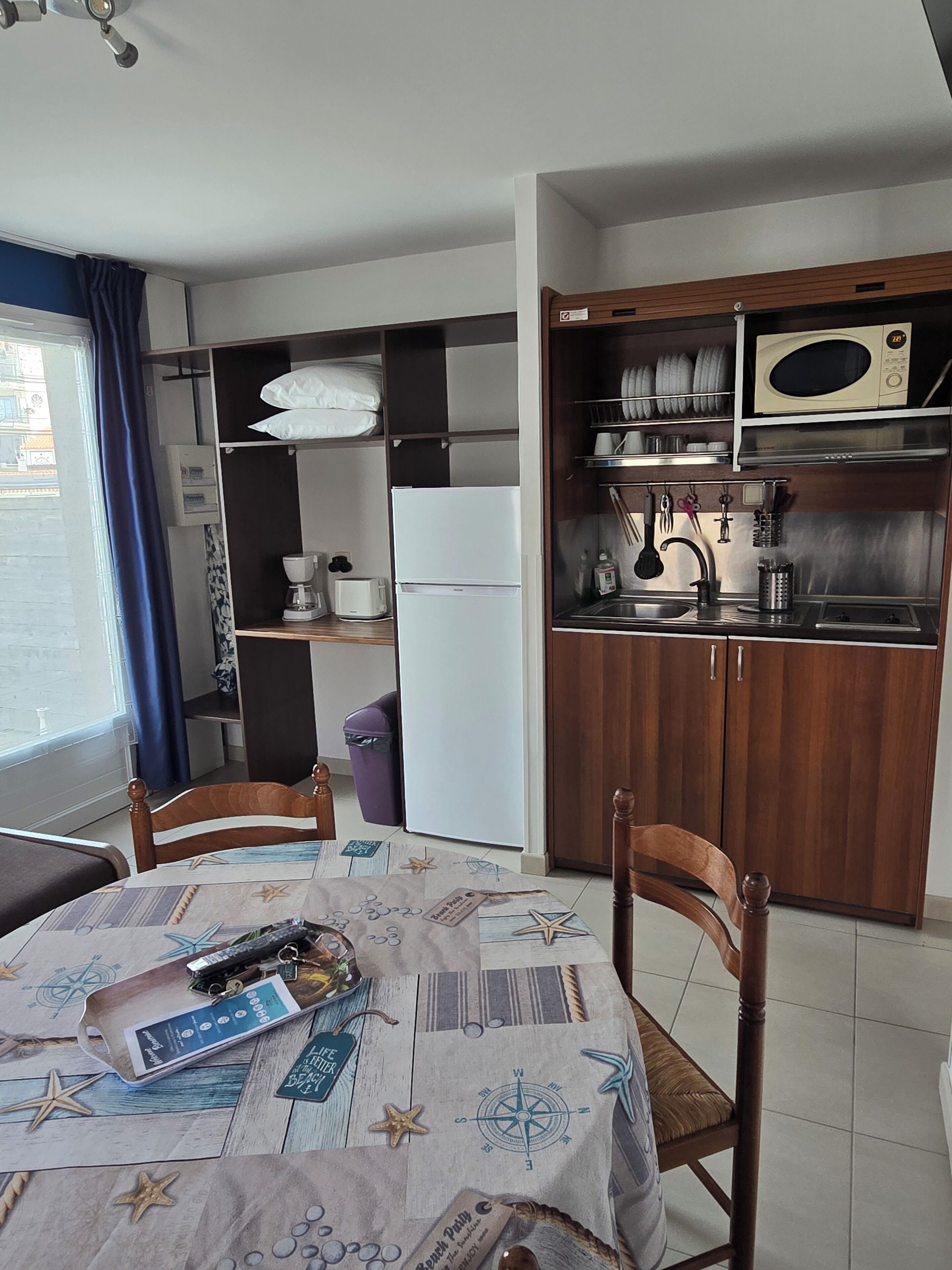 A compact kitchen with a refrigerator, microwave, and sink. There is a dining table with chairs and a window with curtains.