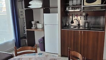 Eine kompakte Küche mit Kühlschrank, Mikrowelle und Spüle. Ein Esstisch mit Stühlen und ein Fenster mit Vorhängen sind vorhanden.