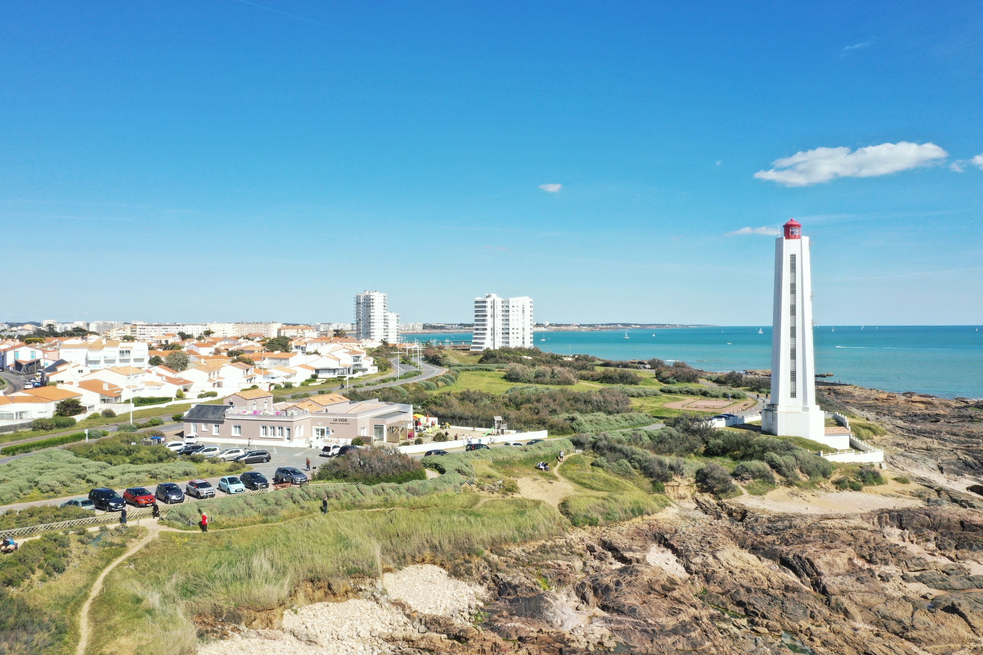 A coastal town with a prominent lighthouse, residential buildings, and a parking area.