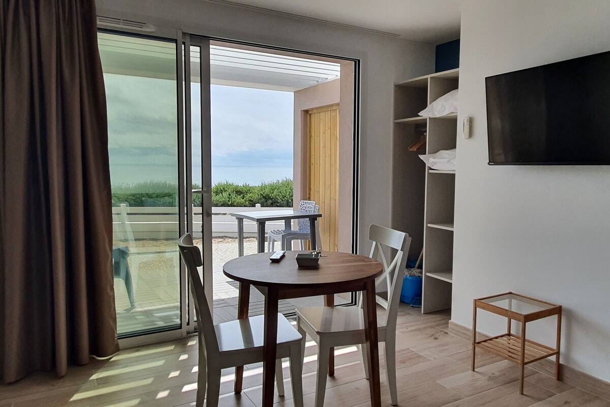 Une pièce dotée d’une porte-fenêtre coulissante donnant sur un espace extérieur, d’une petite table ronde avec des chaises, d’un téléviseur fixé au mur et d’une étagère.