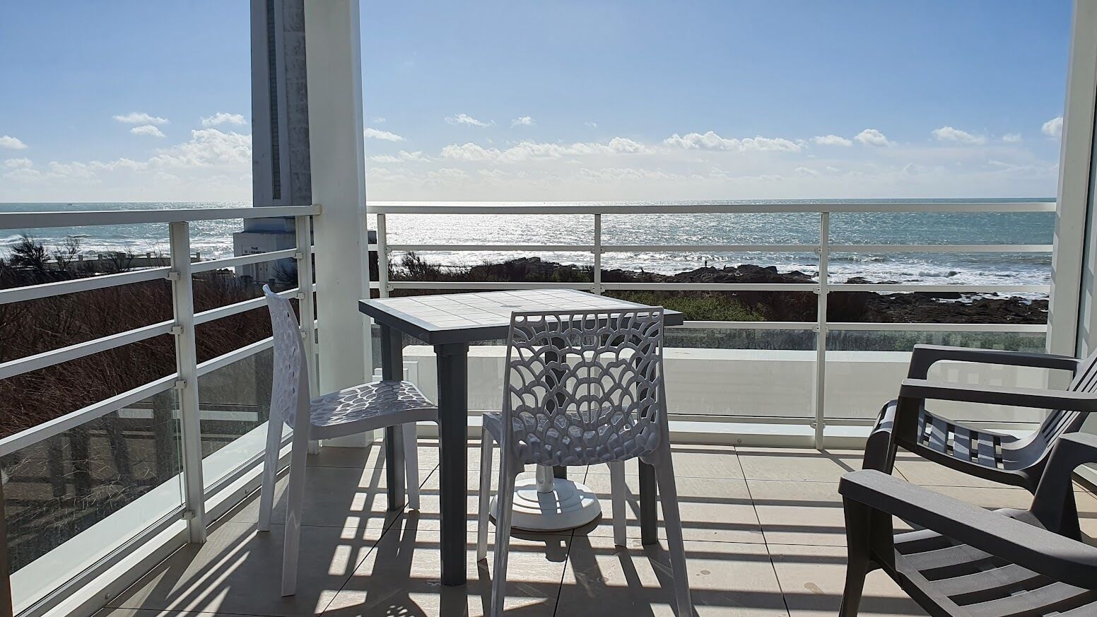 A balcony with a table and chairs, overlooking the ocean with a clear sky.