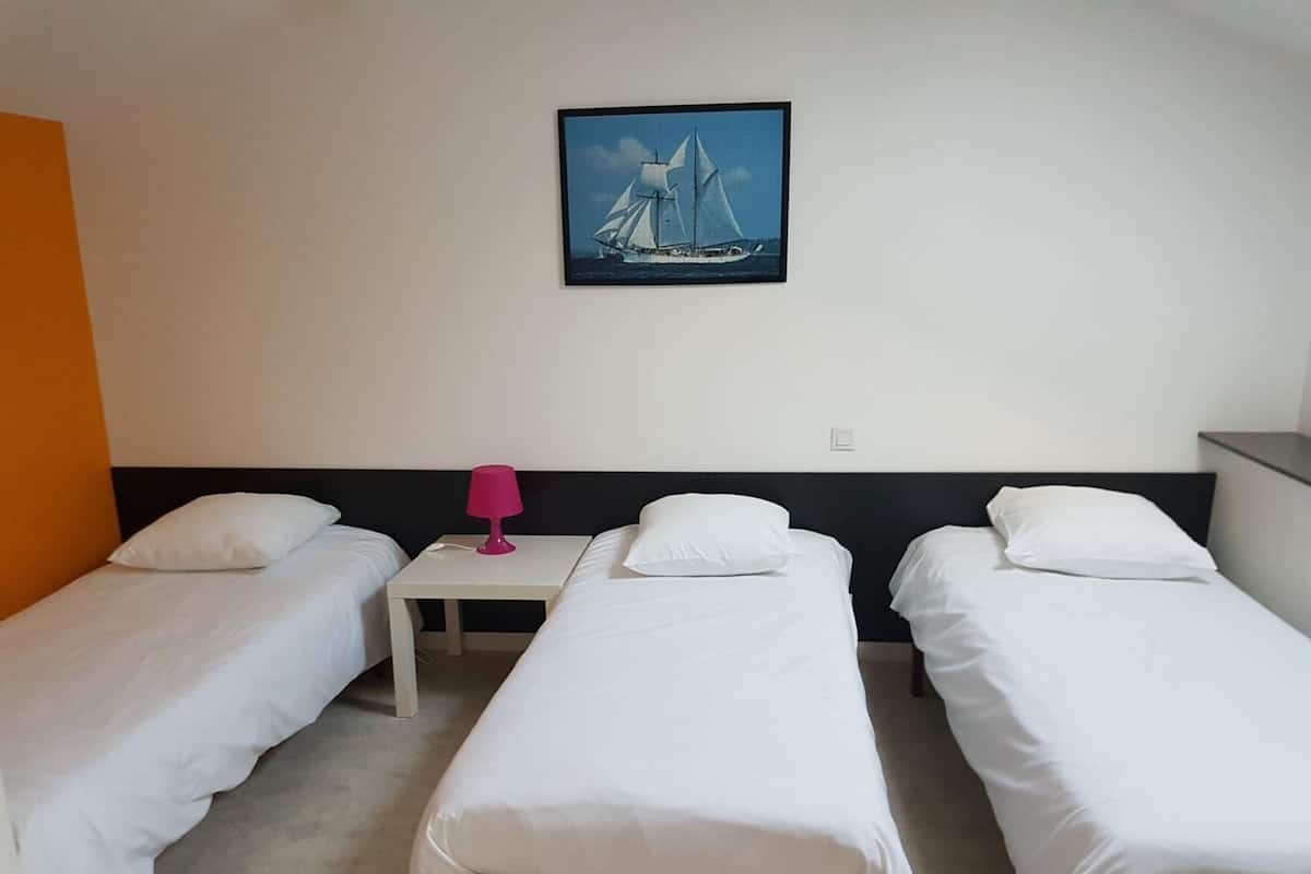 Une chambre d’hôtel avec trois lits simples, une petite table avec une lampe et un tableau encadré au mur.