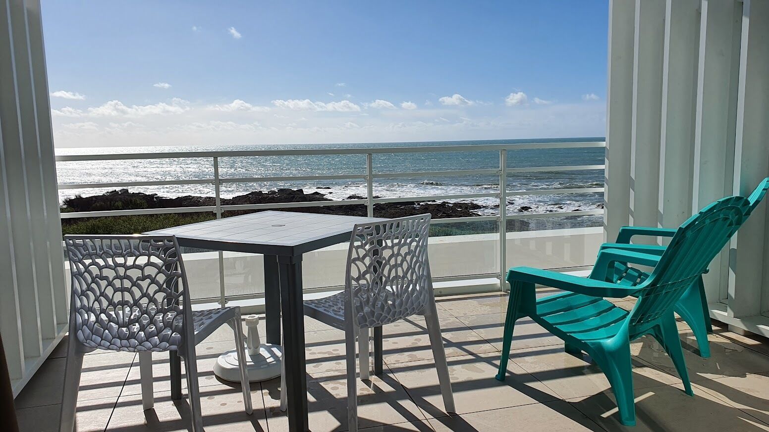 A balcony with a table and chairs, overlooking a rocky coastline and the ocean.