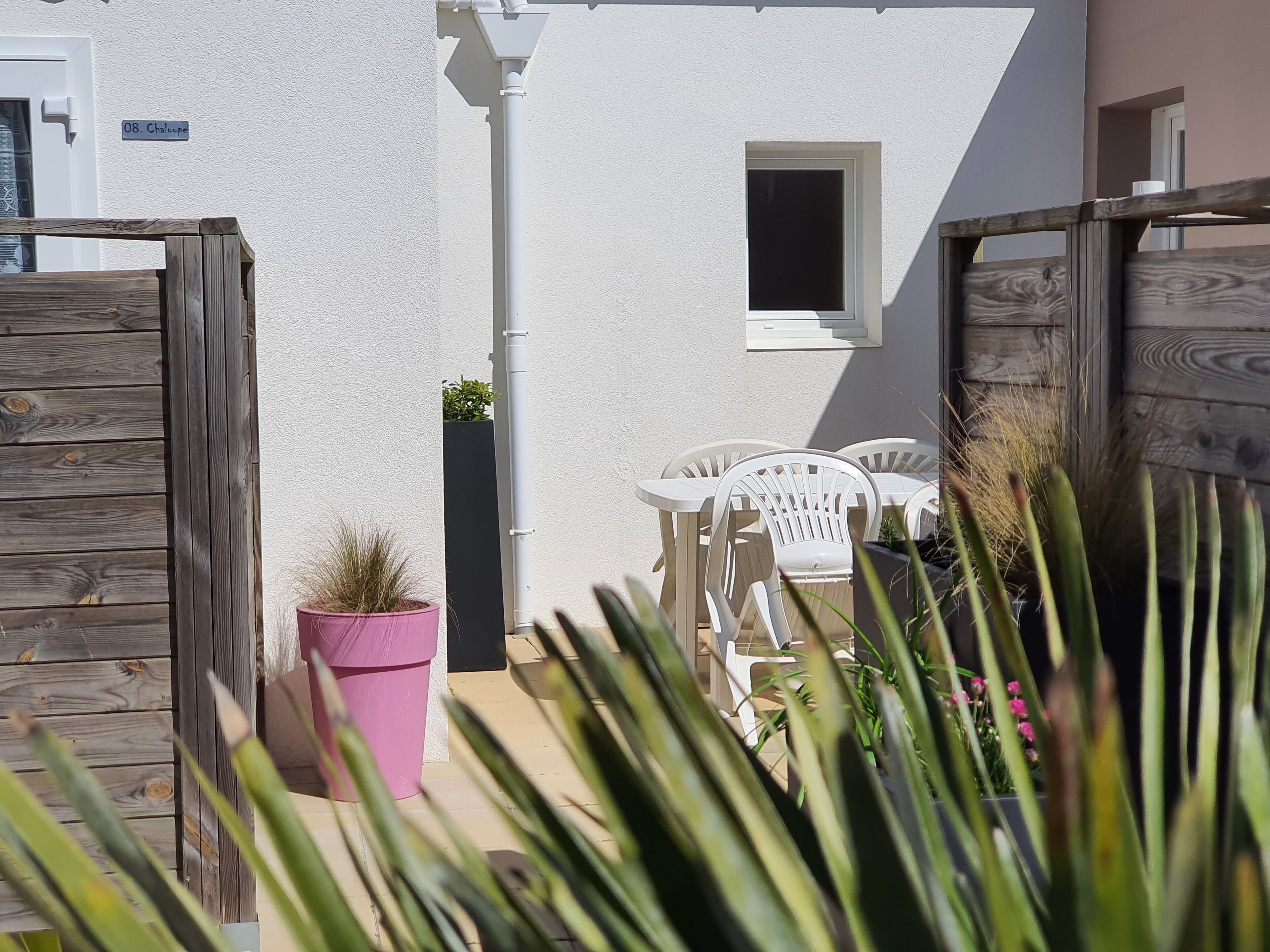 Un bâtiment blanc avec une petite fenêtre, une clôture en bois et une table avec des chaises.